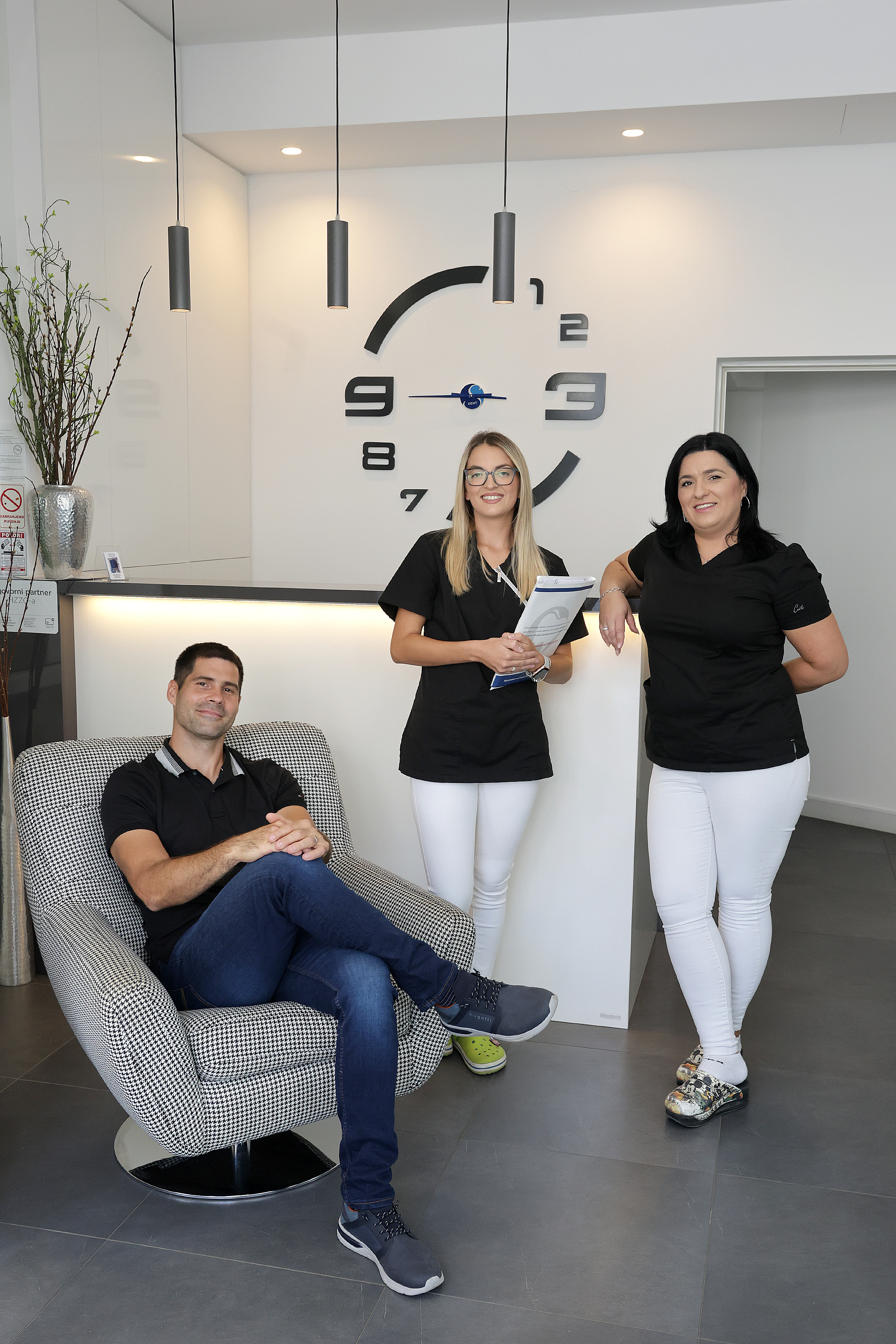 Three people in black tops and white pants posing in a modern reception area with a large wall clock behind them; one man is sitting on a patterned chair and two women are standing.